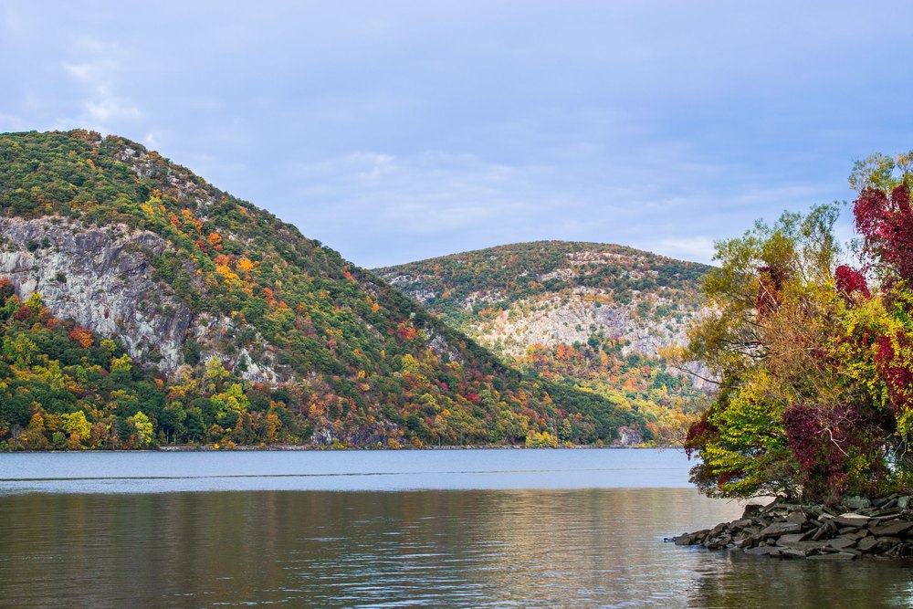 The Hudson River running between low forested hills in full fall color, seen from the bank at Cold Spring